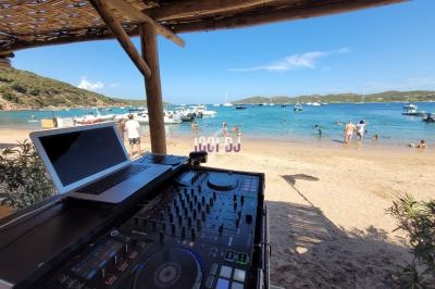 Table de mixage et ordinateur installés sous un abri en bois, avec vue sur une plage animée et des bateaux amarrés.