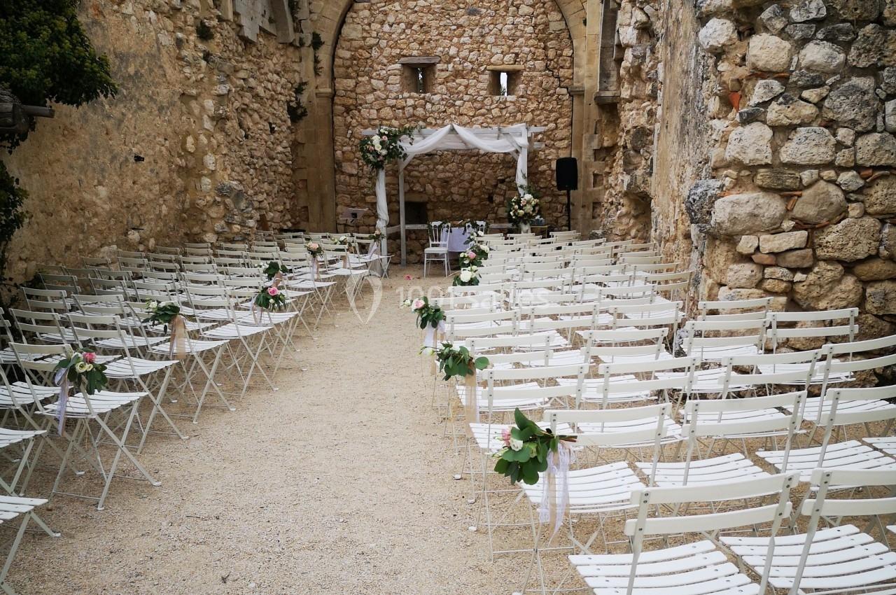 Chaises blanches alignées et décorées, disposées face à une arche ornée de fleurs dans une ruine en pierre.