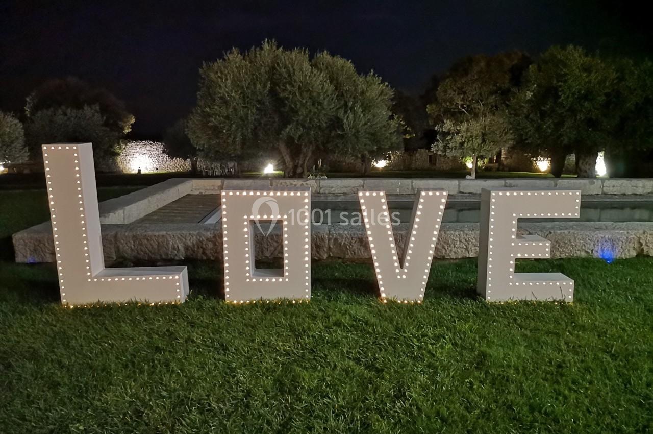 Lettres lumineuses formant le mot ’LOVE’ posées sur une pelouse, avec des arbres et un bassin en arrière-plan.