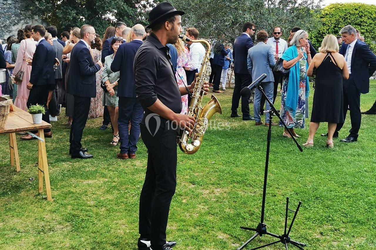 Un musicien joue du saxophone en plein air lors d'un événement avec des invités rassemblés en arrière-plan.