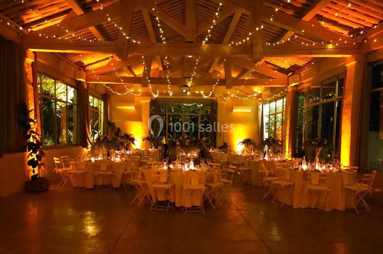 Salle de réception illuminée par des guirlandes lumineuses, avec des tables rondes décorées pour un événement festif.