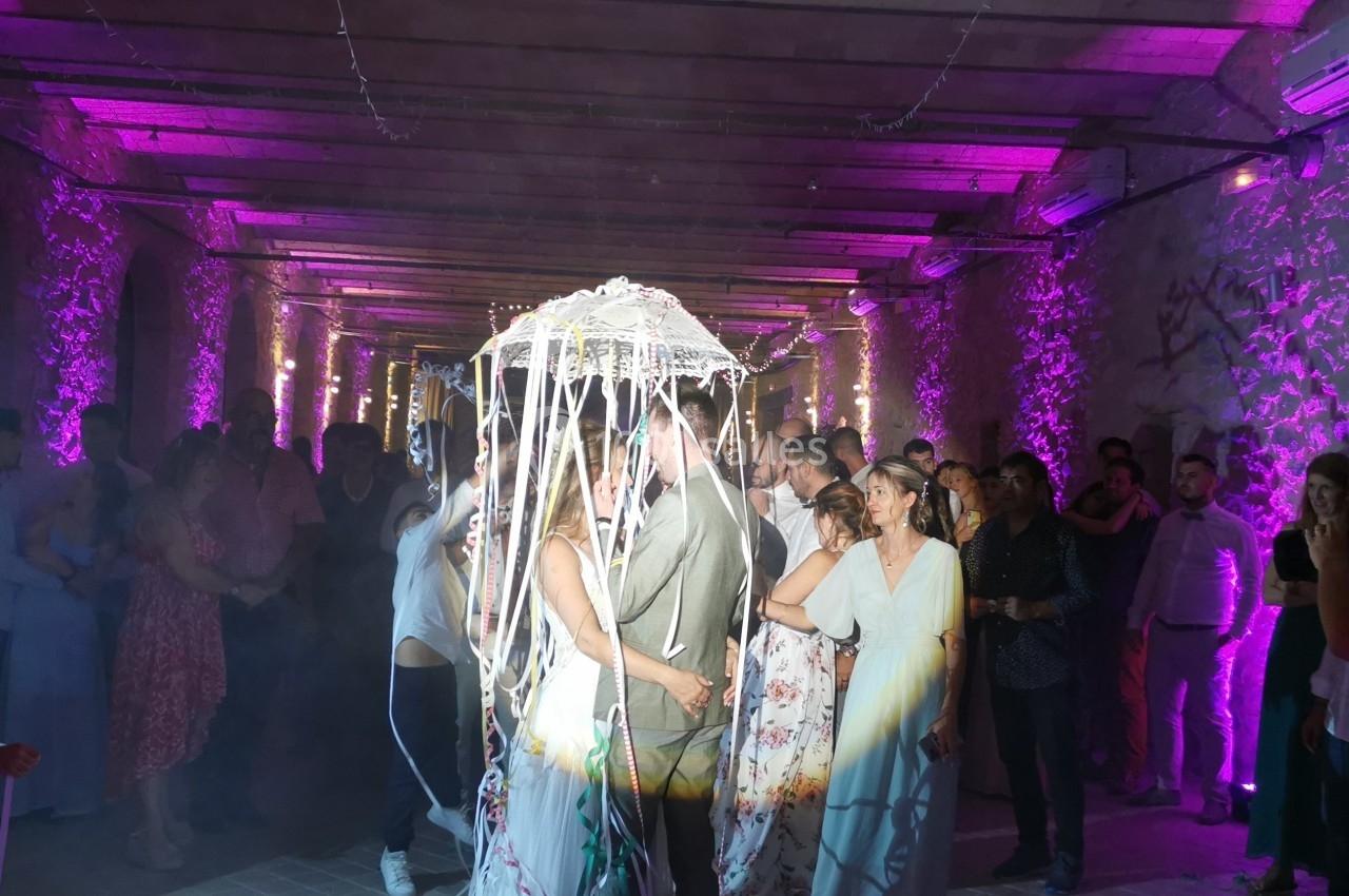 Un couple danse sous une décoration lumineuse en forme de parapluie, entouré d'invités dans une salle éclairée en violet.