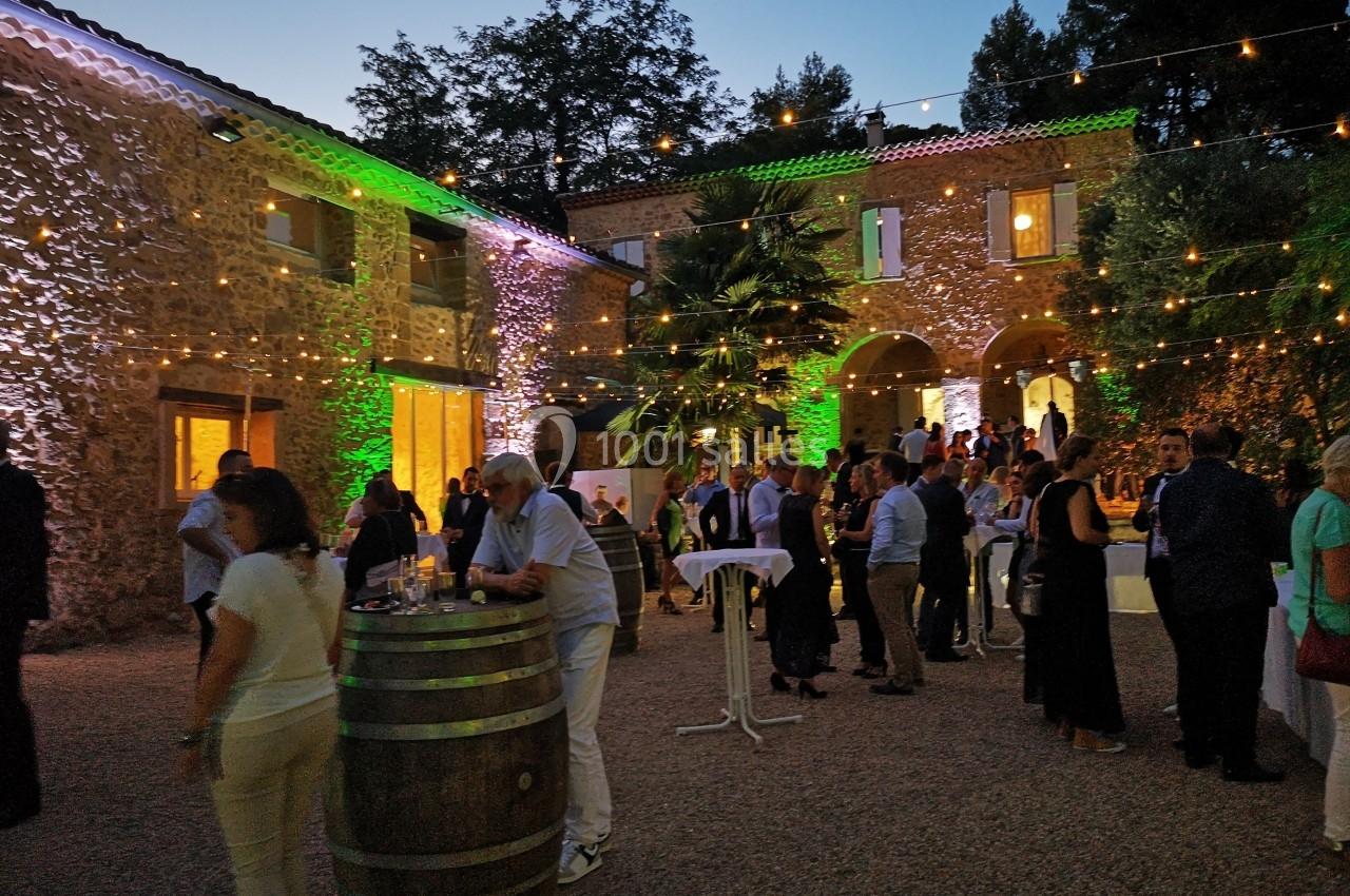 Personnes rassemblées dans une cour en soirée, éclairée par des guirlandes lumineuses et entourée de bâtiments en pierre.