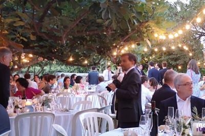 Des convives assis et debout participent à un dîner en extérieur sous des guirlandes lumineuses dans un jardin.
