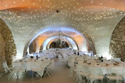 Salle voûtée en pierre décorée de guirlandes lumineuses, tables rondes dressées avec nappes blanches pour un événement.