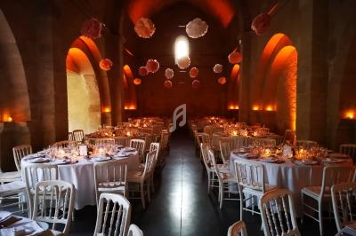 Salle voûtée décorée pour un événement, avec tables rondes dressées, chaises blanches et suspensions florales.
