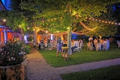 Dîner en plein air sous des guirlandes lumineuses, avec des invités assis à des tables dans un jardin au crépuscule.