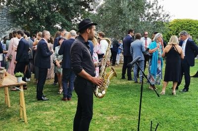 Un musicien jouant du saxophone dans un jardin lors d'un événement en plein air avec des invités en arrière-plan.