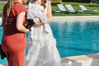 Une femme en robe de mariée sourit près d'une piscine, tenant un bébé en tenue blanche, accompagnée d'une autre femme.