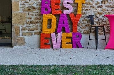 Lettres colorées formant ’Best Day Ever’ appuyées contre un mur en pierre, près d'une table haute rose.