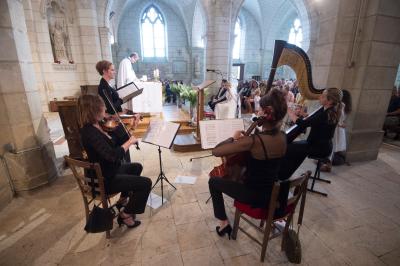 Une violoncelliste joue lors d'une cérémonie dans une église, entourée d'invités debout et de mariés à l'arrière-plan.