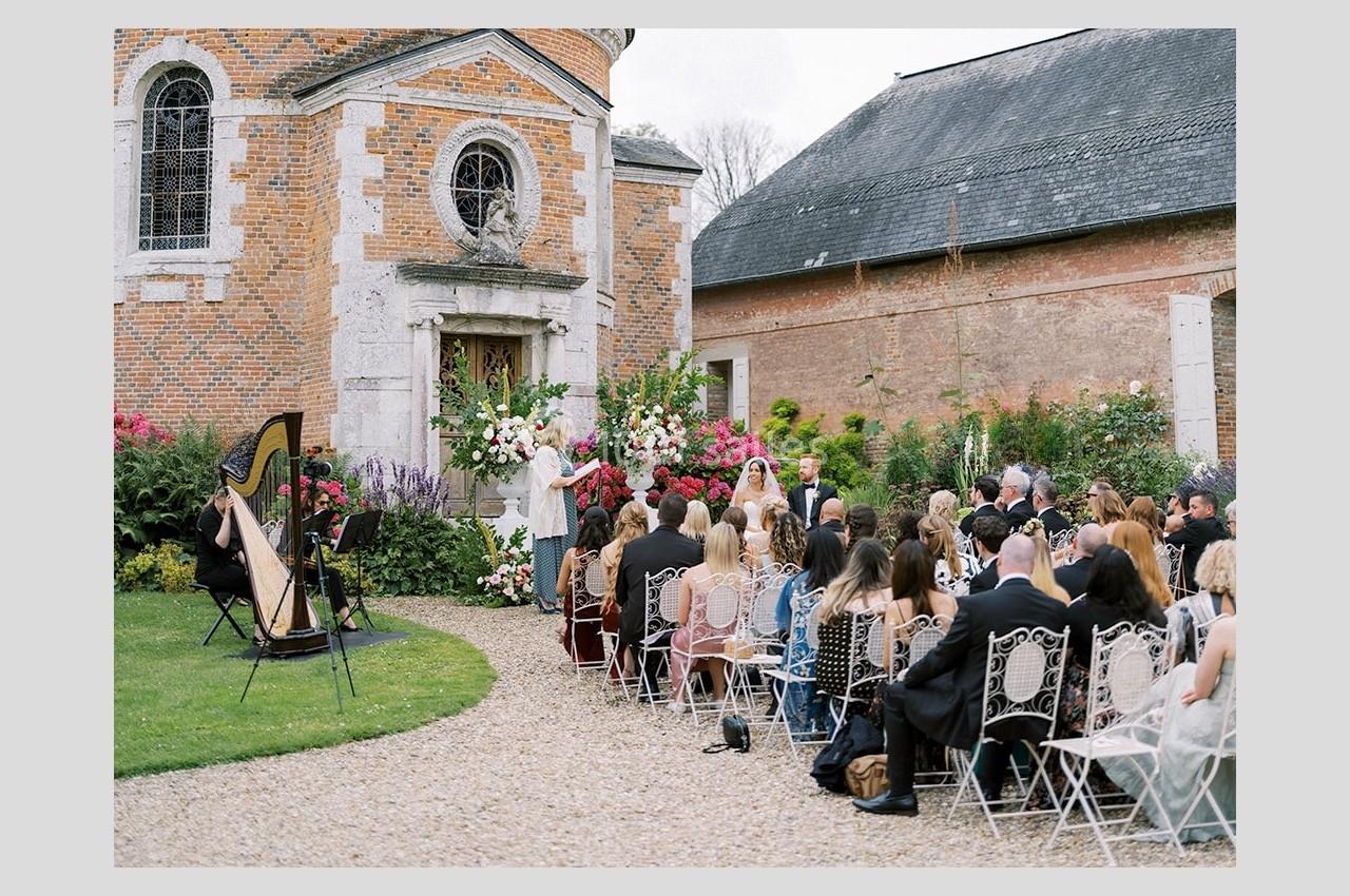 Cérémonie de mariage en plein air devant une chapelle en briques, avec invités assis et une harpiste jouant.