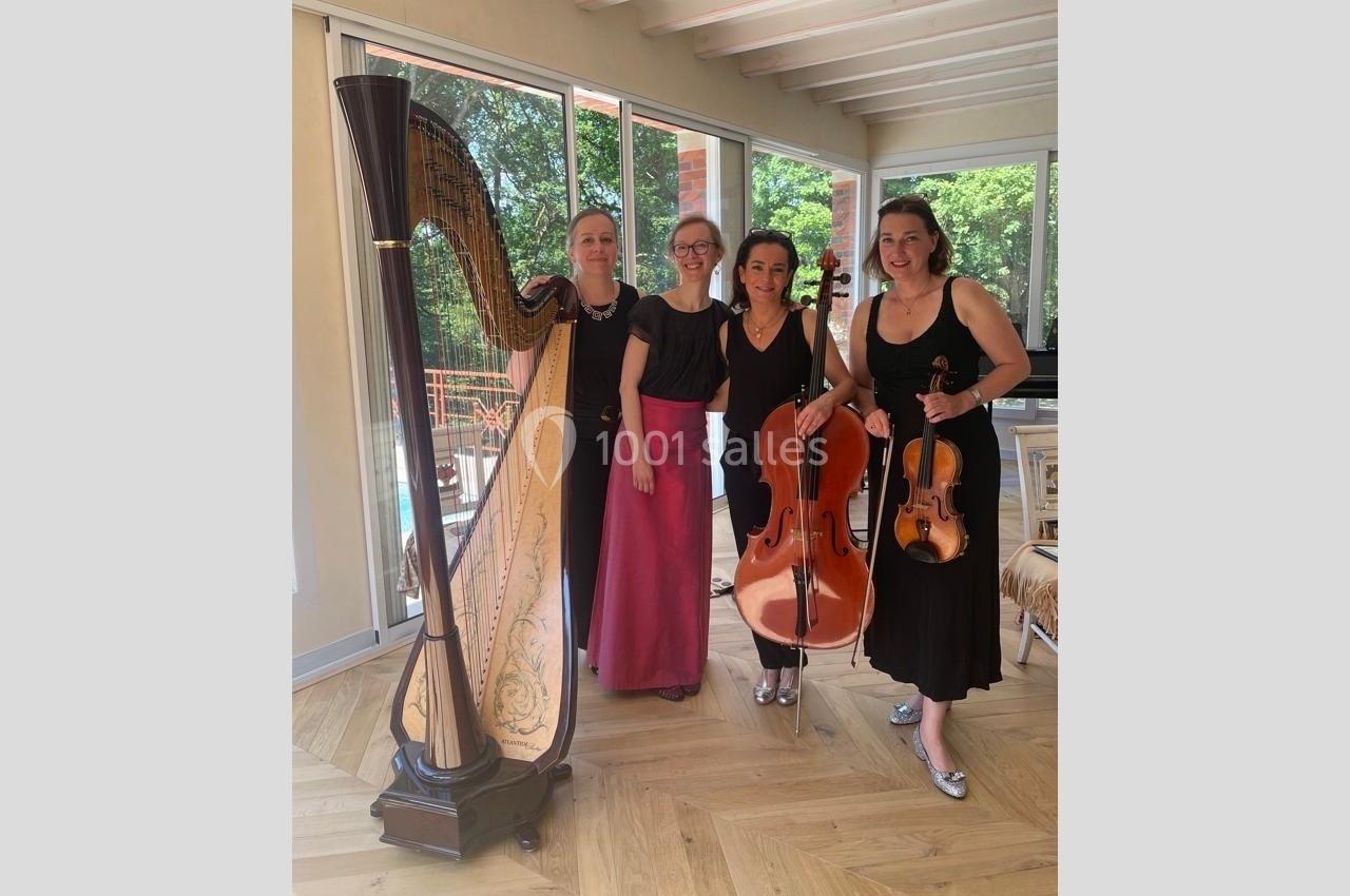 Quatre musiciennes posent avec une harpe, un violoncelle et un violon dans une pièce lumineuse avec parquet en bois.