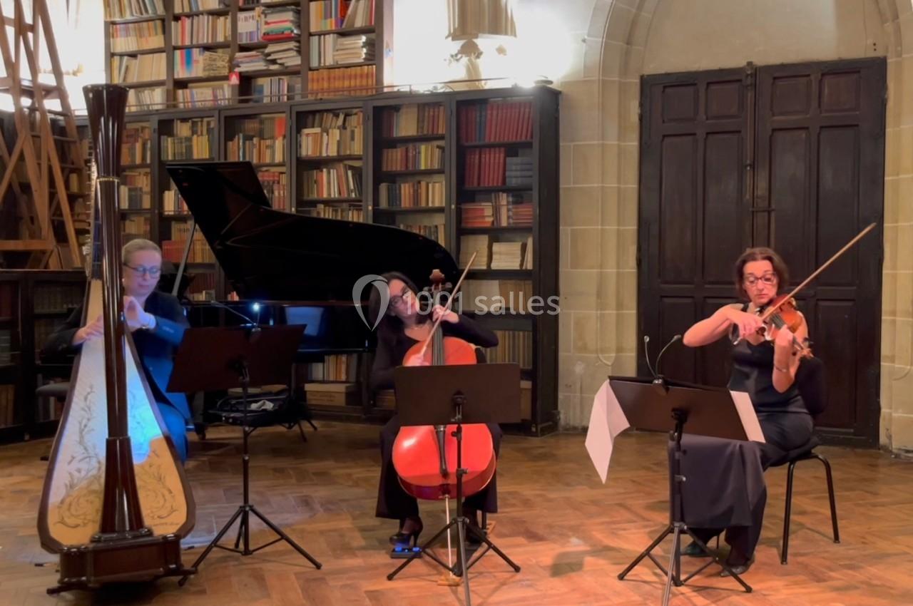 Trois musiciennes jouent de la harpe, du violoncelle et du violon dans une salle avec des étagères remplies de livres.