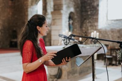 Femme en robe rouge lisant un texte devant un micro dans une église en pierre éclairée par la lumière naturelle.