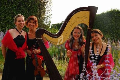 Quatre musiciennes souriantes posent dans un jardin, avec une harpe et un violon au premier plan.