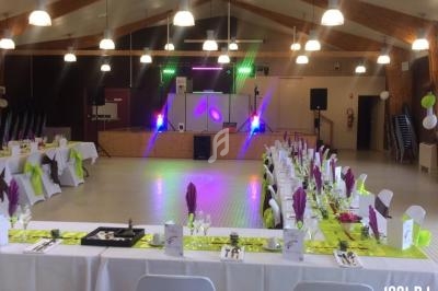 Salle de réception décorée avec des tables dressées en blanc et vert, éclairage coloré sur une scène au fond.