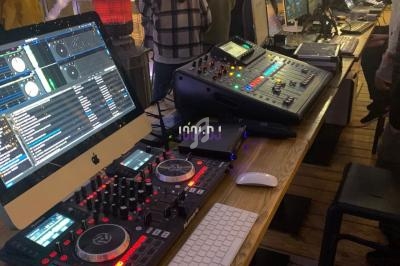 Poste de DJ avec platines, ordinateur, console de mixage et écrans, dans un espace lumineux avec des personnes en arrière…