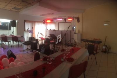 Salle décorée avec des tables dressées, ballons rouges et blancs, et une scène équipée d'instruments de musique.
