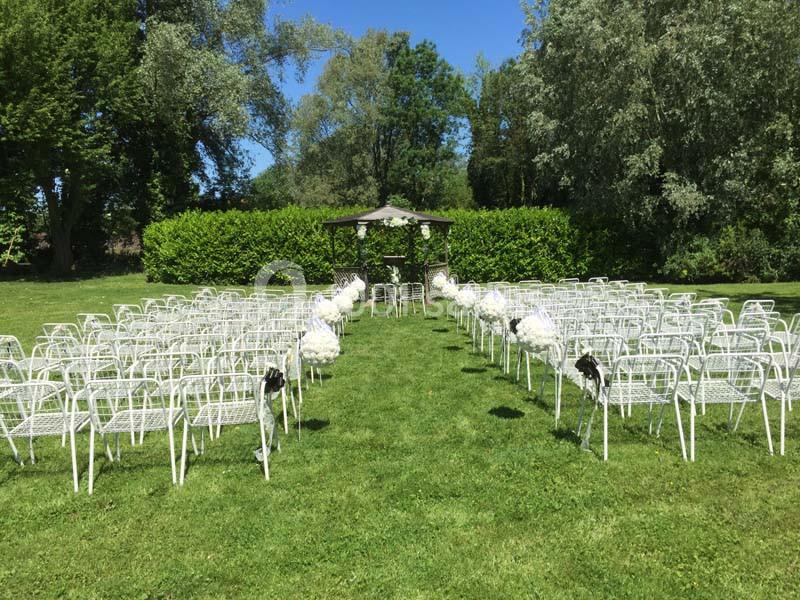 Chaises blanches alignées en extérieur, formant une allée menant à une arche décorée dans un jardin verdoyant.