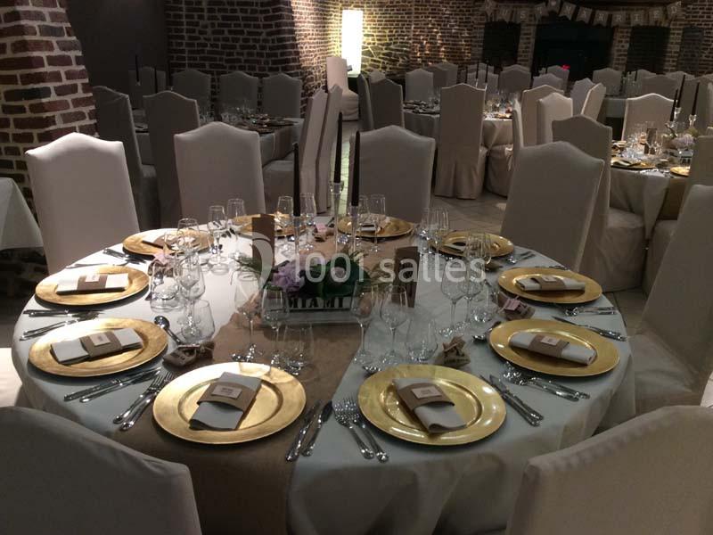 Salle de réception avec tables rondes dressées, nappes beiges, assiettes dorées et décoration florale centrale.