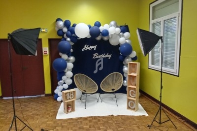 Décor d'anniversaire avec arche de ballons bleus et blancs, deux fauteuils, cubes décoratifs et éclairages de studio.