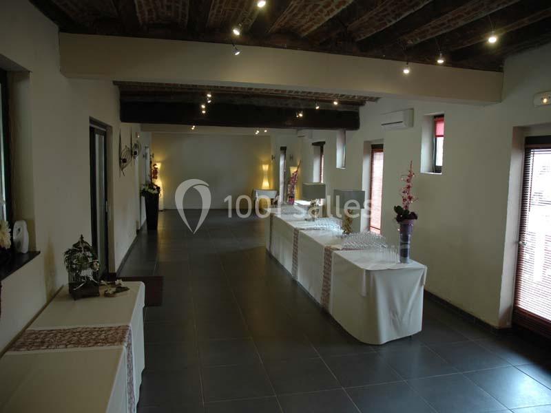 Salle lumineuse avec tables drapées alignées, décorée de plantes et vases, sous un plafond en poutres apparentes.