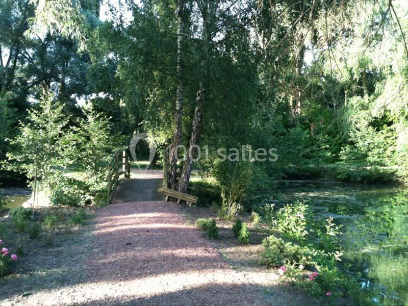 Chemin en gravier bordé de verdure menant à un petit pont en bois au-dessus d'un étang dans un cadre boisé.