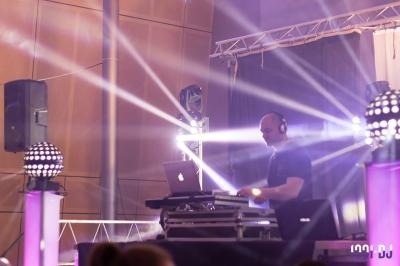 Un DJ mixant sur une scène éclairée par des projecteurs et des faisceaux lumineux dans une salle intérieure.