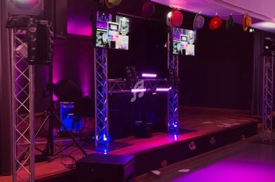 Scène de soirée avec éclairage violet, équipement DJ, écrans et chapeaux colorés suspendus en décoration.