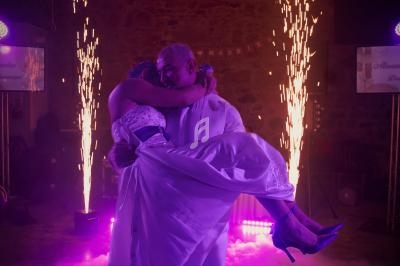 Un homme en costume blanc porte une femme en robe de mariée sur une piste éclairée par des effets lumineux et des étincelles.