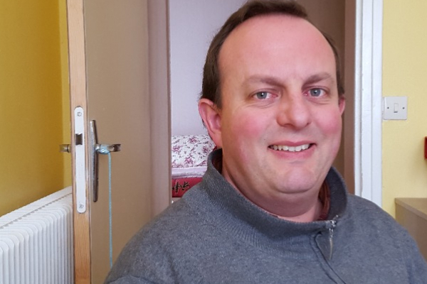 Homme souriant assis dans une pièce avec un mur jaune, une porte ouverte et un lit visible en arrière-plan.