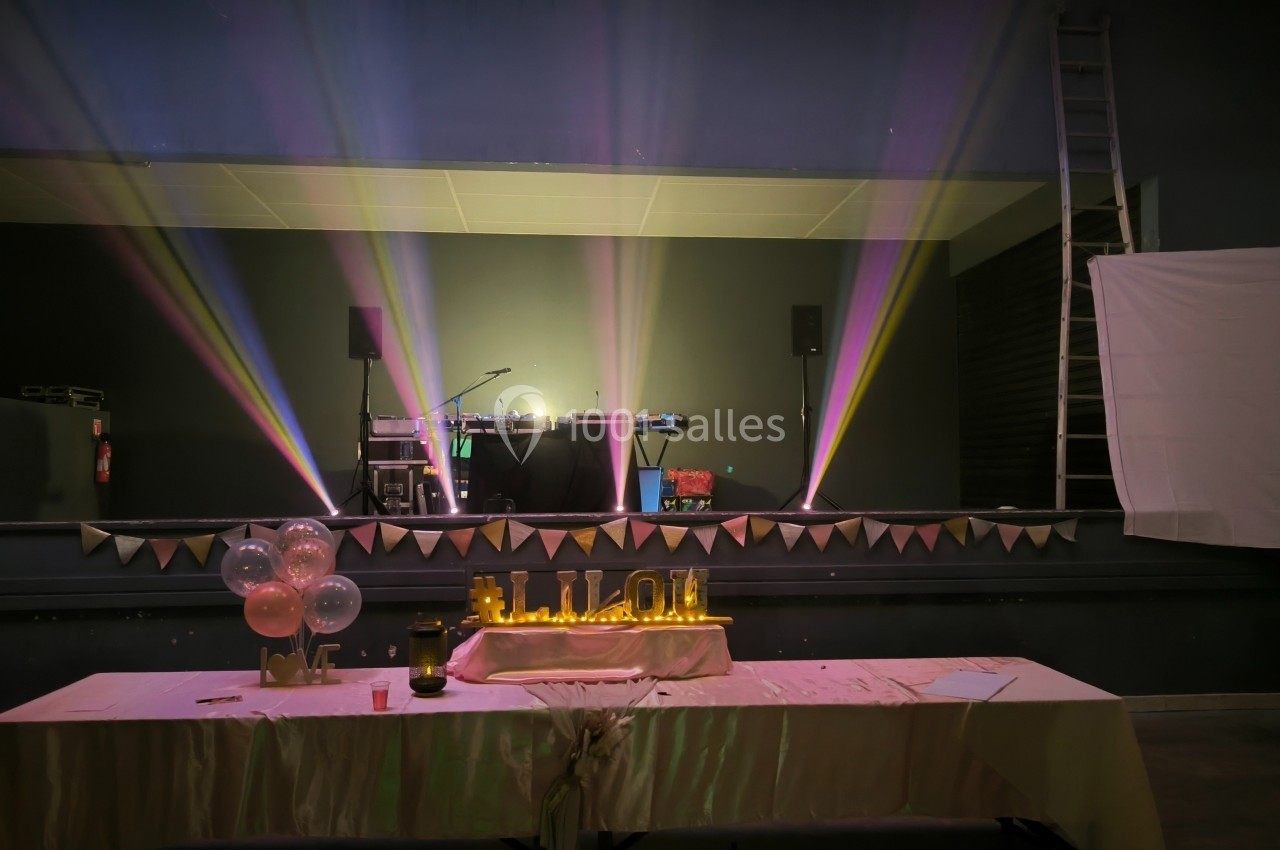 Scène décorée avec des jeux de lumière colorés, une table ornée de ballons et une guirlande lumineuse dorée.