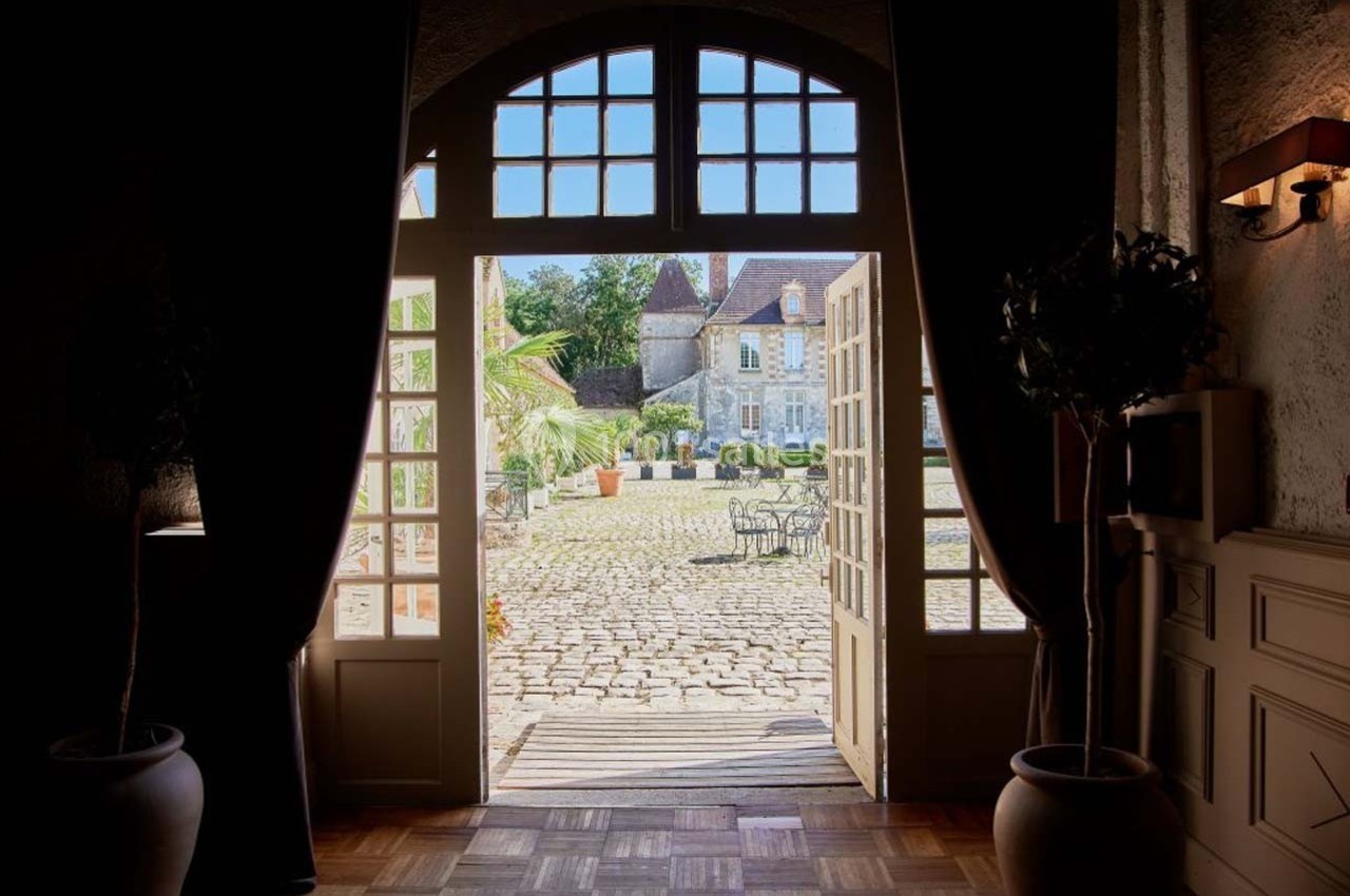 Vue depuis l'intérieur d'une porte ouverte sur une cour pavée ensoleillée avec des tables et un bâtiment ancien en arrière…