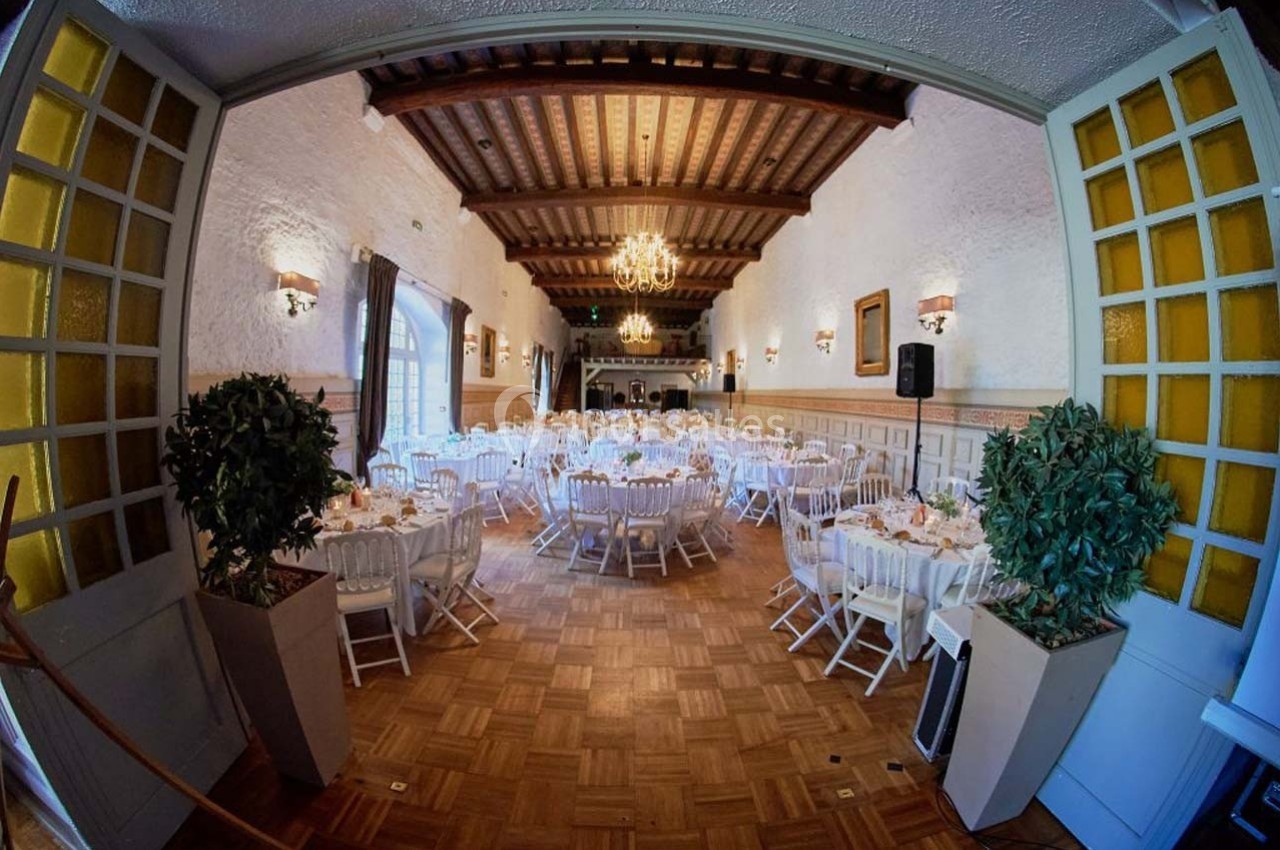 Salle de réception élégante avec tables rondes dressées, chaises blanches et lustres suspendus au plafond en bois.