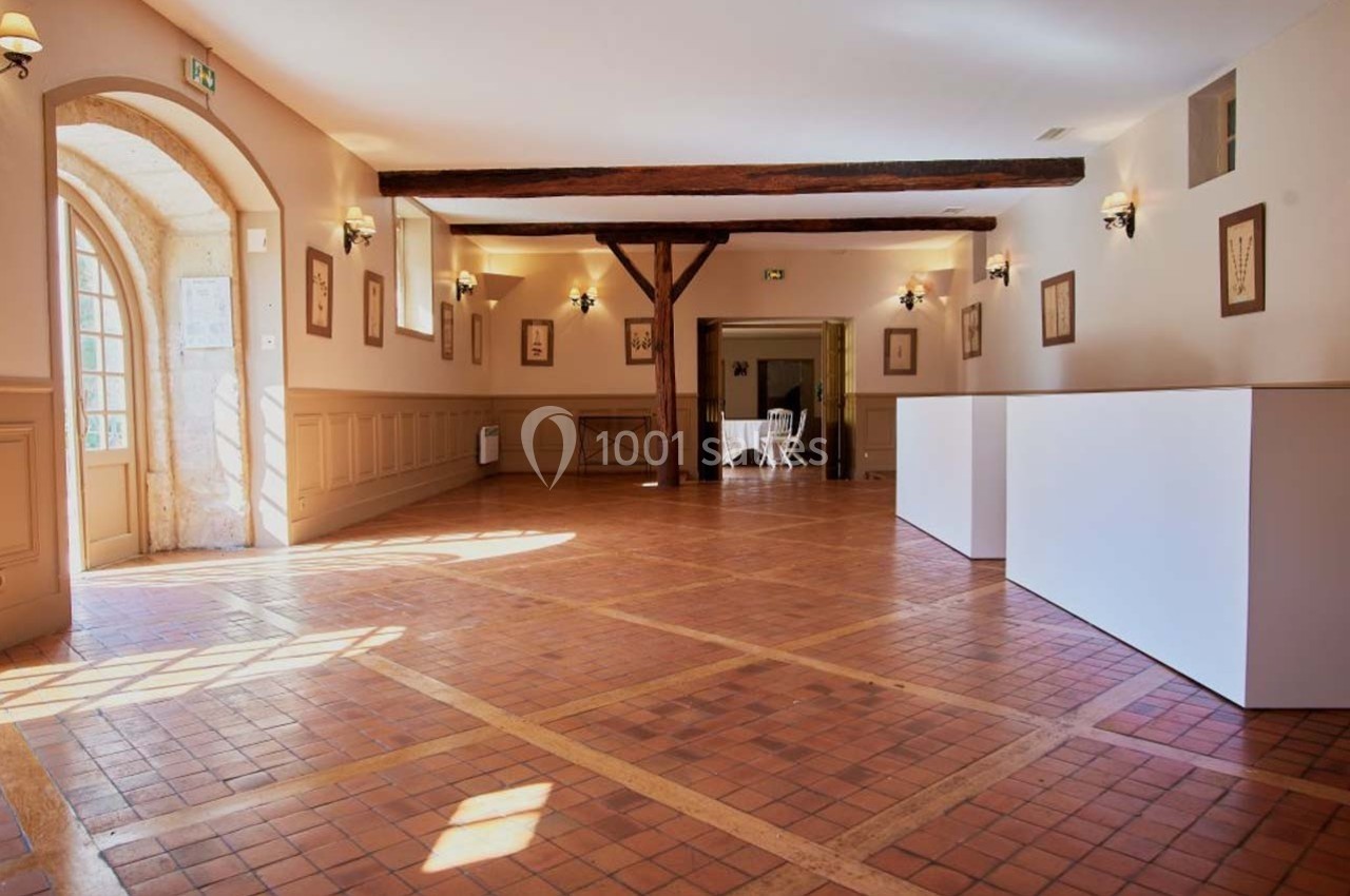 Salle lumineuse avec parquet en terre cuite, poutres apparentes et murs clairs, éclairée par des fenêtres et des appliques.
