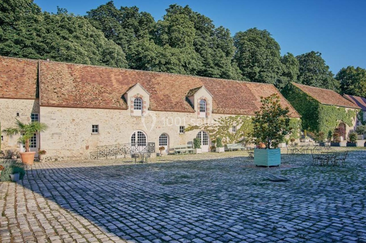 Cour pavée avec bâtiments en pierre, toits en tuiles rouges, entourée de végétation et mobilier de jardin.