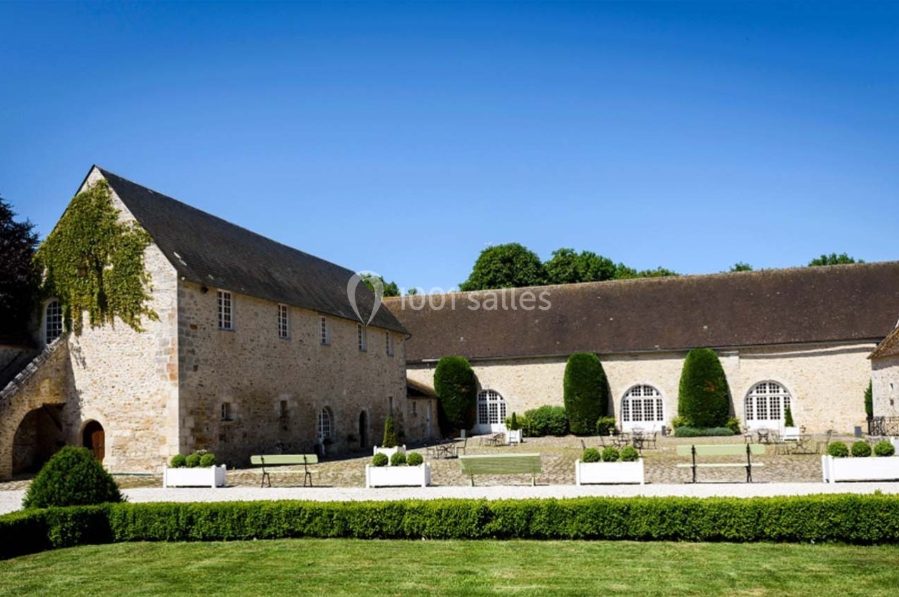 Cour intérieure d'un bâtiment en pierre avec des fenêtres cintrées, des bancs et des haies taillées sous un ciel bleu.