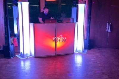 Un DJ mixe derrière une console lumineuse entourée de colonnes éclairées dans une salle au parquet en bois.