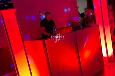 Un DJ mixe derrière une table éclairée de lumières rouges, entouré de colonnes lumineuses et de boules disco.