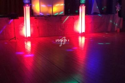 Salle de réception sombre avec éclairages colorés et colonnes lumineuses rouges sur une scène en bois.