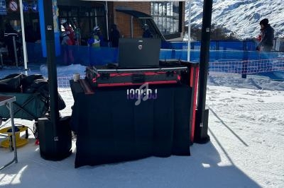 Table de DJ sous une tente blanche en extérieur, installée sur une piste enneigée avec des skieurs en arrière-plan.