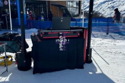Poste de DJ installé sous une tente blanche sur une piste enneigée, avec des skieurs en arrière-plan.