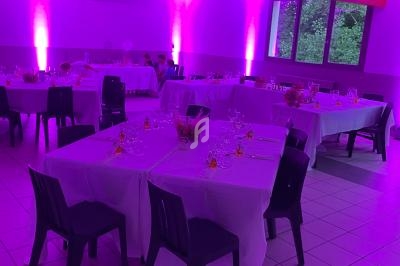 Salle décorée pour un événement, avec tables dressées, nappes blanches et éclairage d'ambiance rose-violet.