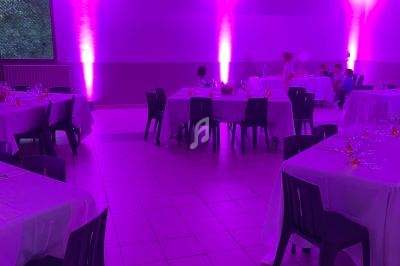 Salle de réception éclairée en rose avec des tables dressées et des chaises noires, quelques personnes en arrière-plan.