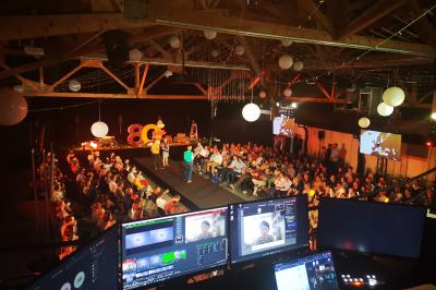 Un artiste sur échasses interagit avec une femme dans un espace intérieur éclairé, avec des spectateurs en arrière-plan.