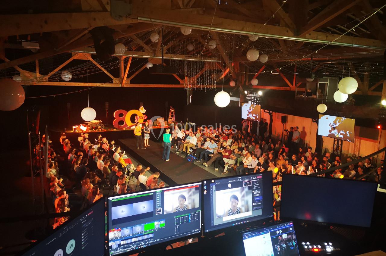 Salle comble avec public assis, scène éclairée, écrans de contrôle au premier plan et décorations suspendues.