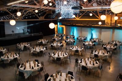 Un artiste sur échasses interagit avec une femme dans un espace intérieur éclairé, avec des spectateurs en arrière-plan.