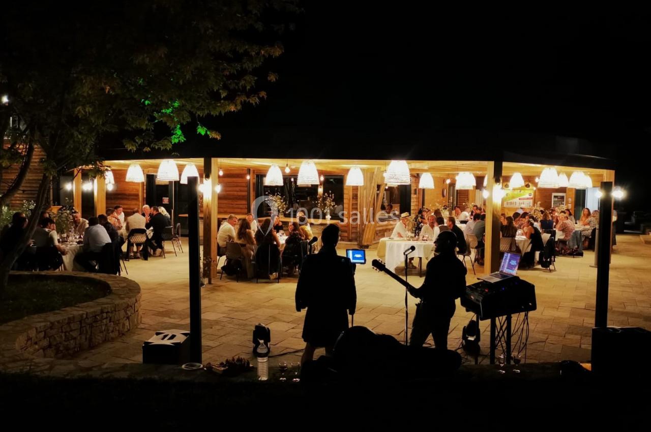 Un groupe de musiciens joue en plein air devant une terrasse éclairée où des convives dînent le soir.