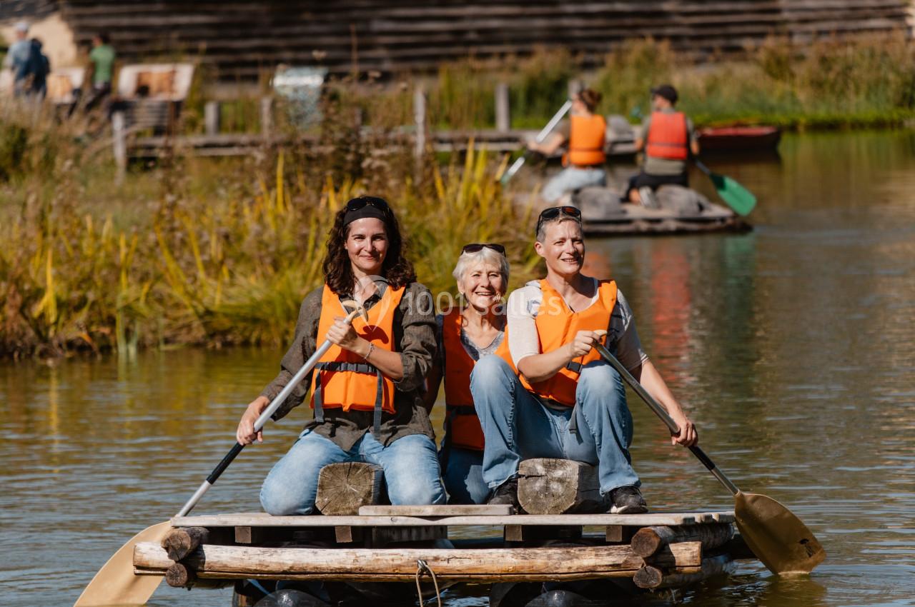 Trois personnes portant des gilets de sauvetage pagaient sur un radeau en bois sur un plan d'eau calme.